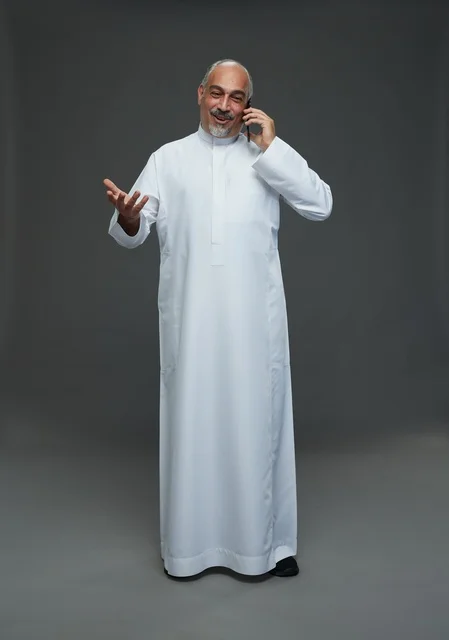 رجل  عربي سعودي خليجي مسن يرتدي الزي الثوب الابيض ويمسك الهاتف المحمول, اجراء مكالمه هاتفيه,  ايماءات واشارات مختلفه , كامل الجسم, خلفيه رماديه