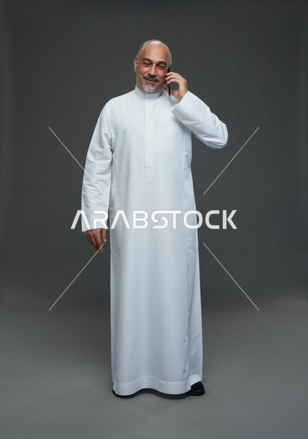 رجل  عربي سعودي خليجي مسن يرتدي الزي الثوب الابيض ويمسك الهاتف المحمول, اجراء مكالمه هاتفيه,  ايماءات واشارات مختلفه , كامل الجسم, خلفيه رماديه
