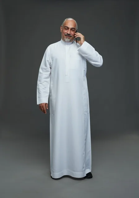 An elderly Arab Saudi Gulf man wearing a white thobe is holding a mobile phone, making a phone call, with various gestures and signals, full body, gray background.