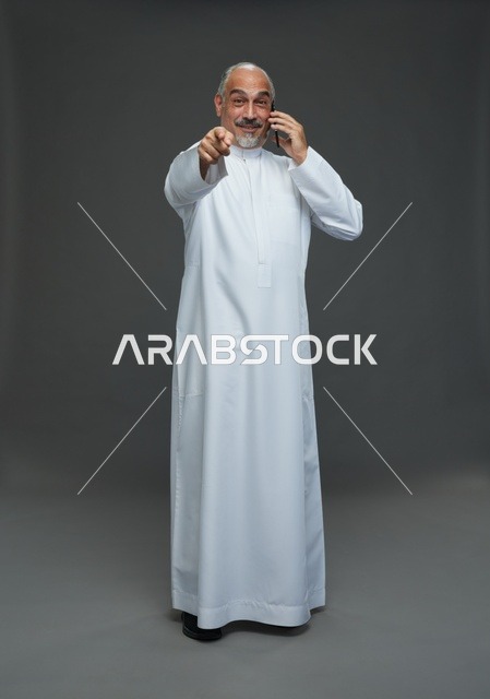 رجل  عربي سعودي خليجي مسن يرتدي الزي الثوب الابيض ويمسك الهاتف المحمول, اجراء مكالمه هاتفيه,  ايماءات واشارات مختلفه , كامل الجسم, خلفيه رماديه