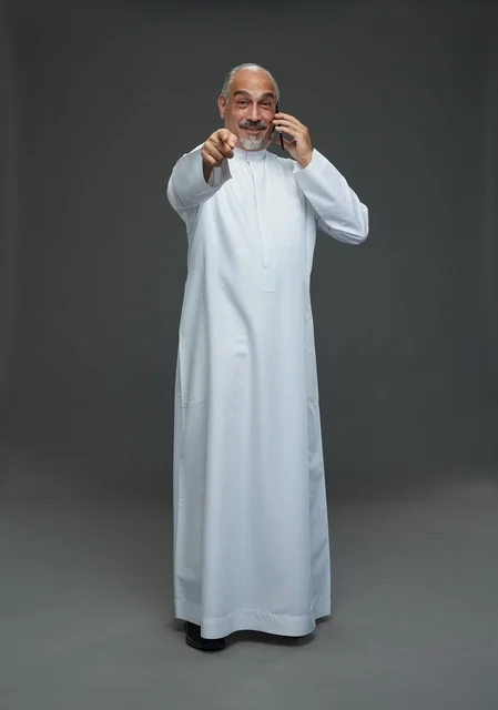 An elderly Arab Saudi Gulf man wearing a white thobe is holding a mobile phone, making a phone call, with various gestures and signals, full body, gray background.