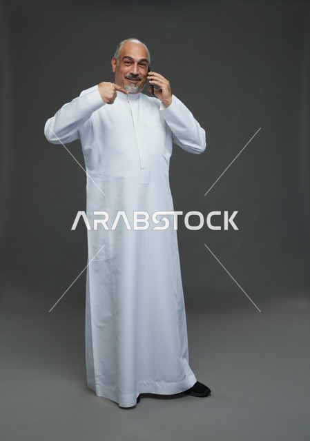 رجل  عربي سعودي خليجي مسن يرتدي الزي الثوب الابيض ويمسك الهاتف المحمول, اجراء مكالمه هاتفيه,  ايماءات واشارات مختلفه , كامل الجسم, خلفيه رماديه
