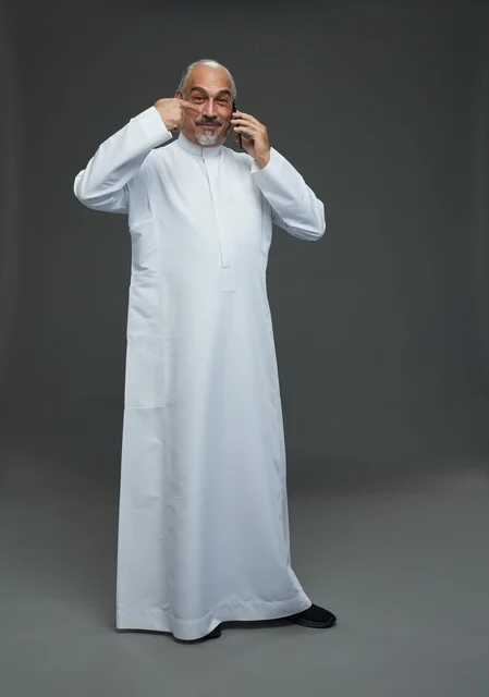 An elderly Arab Saudi Gulf man wearing a white thobe is holding a mobile phone, making a phone call, with various gestures and signals, full body, gray background.
