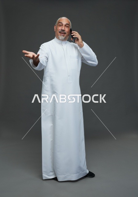 رجل  عربي سعودي خليجي مسن يرتدي الزي الثوب الابيض ويمسك الهاتف المحمول, اجراء مكالمه هاتفيه,  ايماءات واشارات مختلفه , كامل الجسم, خلفيه رماديه