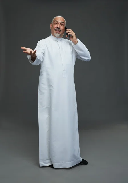 An elderly Arab Saudi Gulf man wearing a white thobe is holding a mobile phone, making a phone call, with various gestures and signals, full body, gray background.