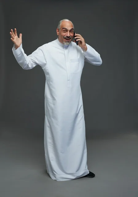 An elderly Arab Saudi Gulf man wearing a white thobe is holding a mobile phone, making a phone call, with various gestures and signals, full body, gray background.