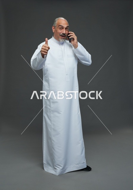 رجل  عربي سعودي خليجي مسن يرتدي الزي الثوب الابيض ويمسك الهاتف المحمول, اجراء مكالمه هاتفيه,  ايماءات واشارات مختلفه , كامل الجسم, خلفيه رماديه