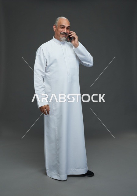 رجل  عربي سعودي خليجي مسن يرتدي الزي الثوب الابيض ويمسك الهاتف المحمول, اجراء مكالمه هاتفيه,  ايماءات واشارات مختلفه , كامل الجسم, خلفيه رماديه