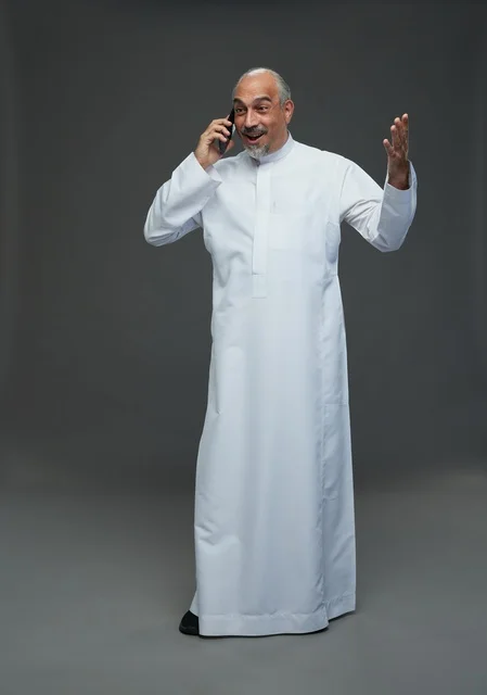 An elderly Arab Saudi Gulf man wearing a white thobe is holding a mobile phone, making a phone call, with various gestures and signals, full body, gray background.