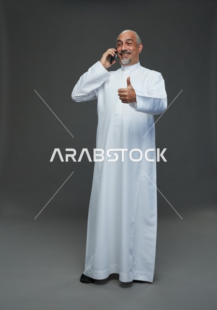 رجل  عربي سعودي خليجي مسن يرتدي الزي الثوب الابيض ويمسك الهاتف المحمول, اجراء مكالمه هاتفيه,  ايماءات واشارات مختلفه , كامل الجسم, خلفيه رماديه