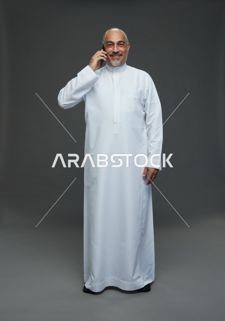 رجل  عربي سعودي خليجي مسن يرتدي الزي الثوب الابيض ويمسك الهاتف المحمول, اجراء مكالمه هاتفيه,  ايماءات واشارات مختلفه , كامل الجسم, خلفيه رماديه