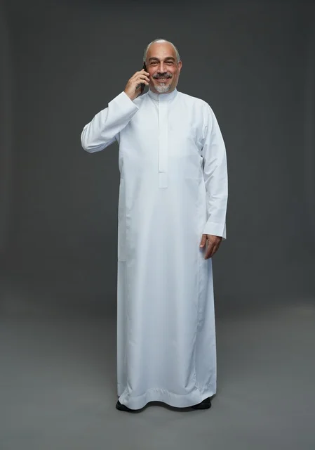 An elderly Arab Saudi Gulf man wearing a white thobe is holding a mobile phone, making a phone call, with various gestures and signals, full body, gray background.