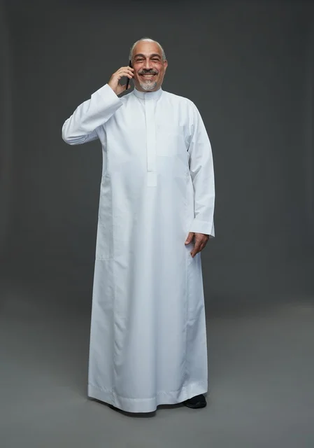 An elderly Arab Saudi Gulf man wearing a white thobe is holding a mobile phone, making a phone call, with various gestures and signals, full body, gray background.
