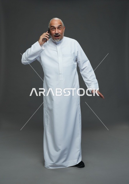 An elderly Arab Saudi Gulf man wearing a white thobe is holding a mobile phone, making a phone call, with various gestures and signals, full body, gray background.