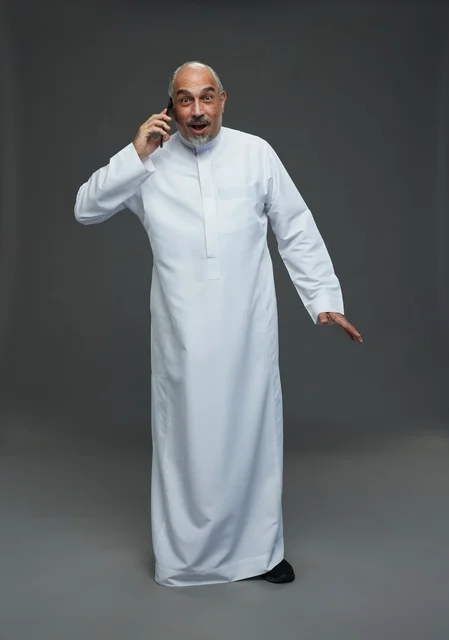An elderly Arab Saudi Gulf man wearing a white thobe is holding a mobile phone, making a phone call, with various gestures and signals, full body, gray background.