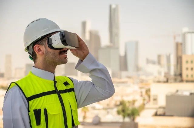  الوظائف الشبابية في المملكة السعودية، نمو وتطور الهندسة المعمارية على ايدي المبدعين أبناء الوطن، صورة جانبية لمهندس عربي خليجي سعودي يرتدي الثوب التقليدي والسترة الخاصة وخوذة الحماية ينظر في نظارة واقع افتراضي ثلاثية الأبعاد، خلفية أبراج وناطحات سحاب 