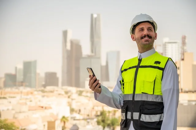 ززThe development and growth of the engineering sector in the Kingdom, construction by the hands of the sons of the nation, a Saudi Arabian Gulf engineer wearing the traditional dress, a special jacket and a protective helmet stands and holds a mobile phone, following up on work according to the workflow strategy, the concept of engineering and architectural construction, a background of towers and skyscrapers
