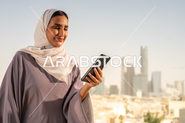تصفح الانترنت و مواقع التواصل الاجتماعي عبر جهاز الجوال،سيدة عربية خليجية سعودية ترتدي العباءة تقوم باستخدام الهاتف المحمول بايماءات السعادة والسرور،الخروج في الهواء الطلق،التواصل مع الاهل و الاصدقاء
