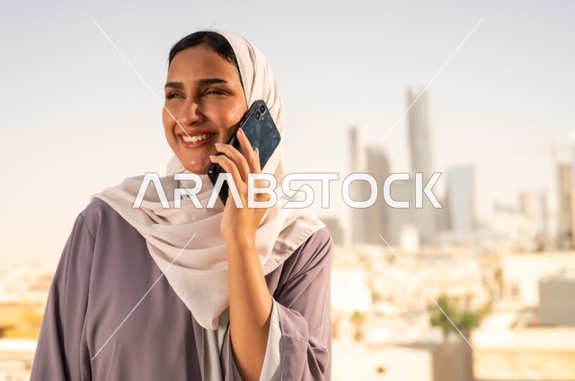 Making a phone call via a mobile device, a close-up image of a Gulf Arab Saudi woman wearing an abaya using a mobile phone with gestures of happiness and joy, going outdoors, communicating with family and friends.