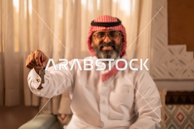 A close-up image of an Arab Gulf man sitting inside a traditional council with Arab decorations and traditional ornaments, holding a house key in his hand, wearing glasses, representing ownership, security, and traditional heritage, creating a beautiful balance between Arab authenticity and beauty, dressed in a white thobe, a red ghutrah, and an agal.