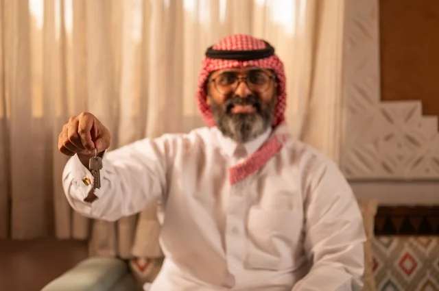 صورة مقربة لرجل عربي خليجي يجلس داخل مجلس تراثي بديكورات عربية وزخارف تقليدية يمسك بيده مفتاح منزل، يضع نظارة طبية، الملكية والأمان والحياة التقليدية، توازناً جميلاً بين الأصالة العربية والجمال، يرتدي الثوب الأبيض والشماغ الأحمر والعقال
