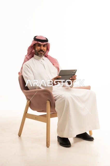 Chatting with family and friends, using modern technology and techniques, a portrait of an Arab Gulf Omani man wearing traditional thobe and shmagh holding a tablet in his hand, browsing social media, using applications and programs on tablet devices, white background.
