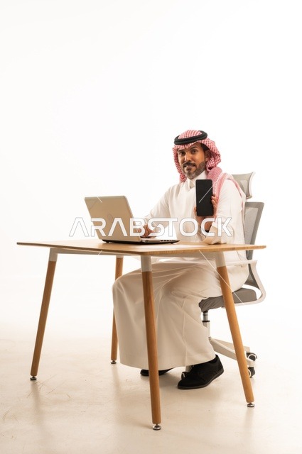 Displaying a blank black screen on the phone, completing work tasks online, using modern technology and techniques, a portrait of an Arab Gulf Omani man wearing traditional attire sitting behind holding a laptop, browsing social media, using mobile applications, white background.