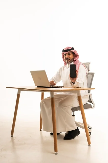 Displaying a blank black screen on the phone, completing work tasks online, using modern technology and techniques, a portrait of an Arab Gulf Omani man wearing traditional attire sitting behind holding a laptop, browsing social media, using mobile applications, white background.