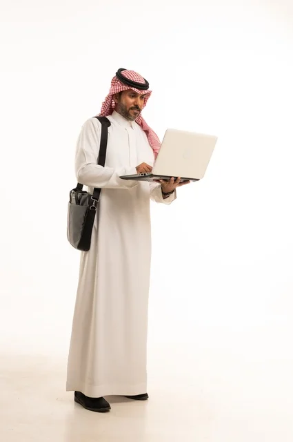 Chatting and conversations and social communication, conducting a business meeting online, completing remote work tasks, online shopping and online orders, a portrait of an Arab Saudi Gulf man wearing traditional thobe and ghutrah, carrying a briefcase and using a laptop, full body, white background.