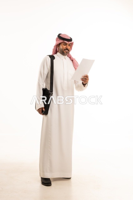 قراءة وتدقيق الملاحظات، مفهوم إداة الأعمال والمشاريع، الاستعداد للذهاب إلى العمل، بورتريه لرجل عربي خليجي سعودي يرتدي الثوب التقليدي والشماغ يحمل بيده حقيبة يد وورقة بيضاء، الأناقة والاهتمام بالمظهر الخارجي، الجسم بالطول الكامل، خلفيه بيضاء