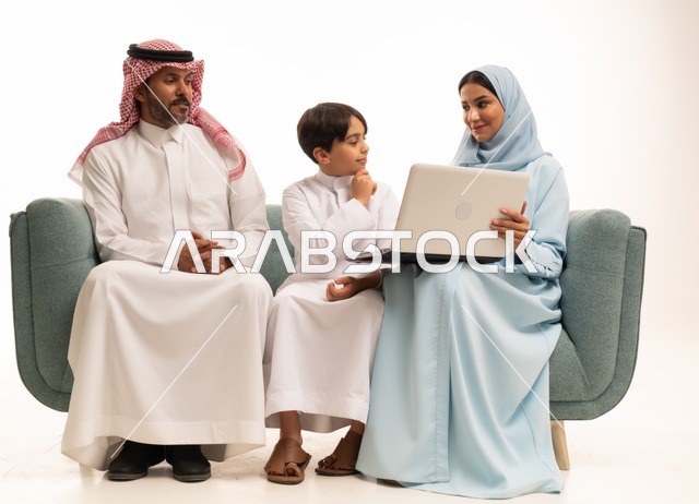متابعة حصص الأونلاين وإجراء مكالمة فيديو من خلال الحاسوب المحمول، استخدام جهاز تقني حديث، بورتريه لولد عربي سعودي خليجي يرتدي الثوب التقليدي يحمل الحاسوب ويجلس على الاريكة برفقة والديه، الأجواء العائلية المليئة بالحب والترابط والإستقرار، خلفية بيضاء