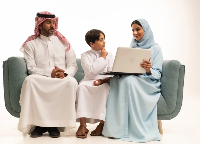 Following online classes and making a video call through a laptop, using modern technology, a portrait of an Arab Saudi Gulf boy wearing traditional attire holding a laptop and sitting on the couch with his parents, a family atmosphere filled with love, connection, and stability, white background.