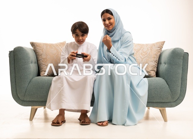 مفهوم الأسرة السعيدة، بورتريه لسيدة عربية خليجية سعودية تجلس في غرفة المعيشة برفقة ابنها، قضاء وقت ممتع بلعب ألعاب الفيديو بإيماءات تدل على السعادة ، ولد يمسك عصا التحكم وتلعب بألعاب الفيديو بجانب والدته ، المتعة و التسلية  بألعاب البلايستيشن الترفيهية ، خلفية بيضاء