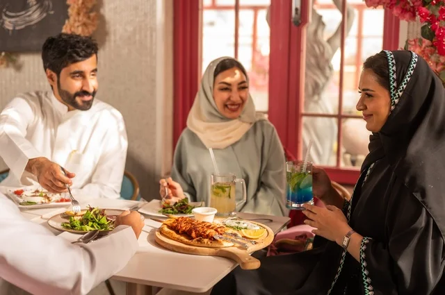 Enjoying lunch at the restaurant, a fun and social gathering, building successful social relationships, exchanging conversations and news, sharing enjoyable moments with friends, a family of Gulf Arab Saudis sitting at the table in one of the upscale restaurants, eating with gestures of happiness and joy.
