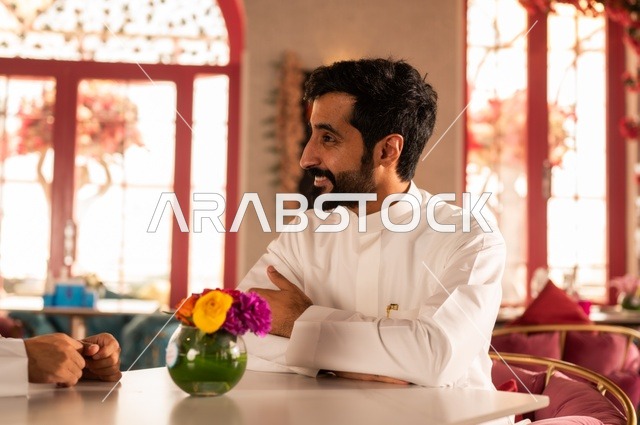 صورة جانبية لشاب  في كوفي شوب راقي، قضاء وقت الفراغ باحتساء المشروب المفضل،  الاستمتاع بالمشروبات الباردة، رجل عربي خليجي سعودي يرتدي الثوب الأبيض يجلس في مقهى على طاولة ، إيماءات السعادة والسرور