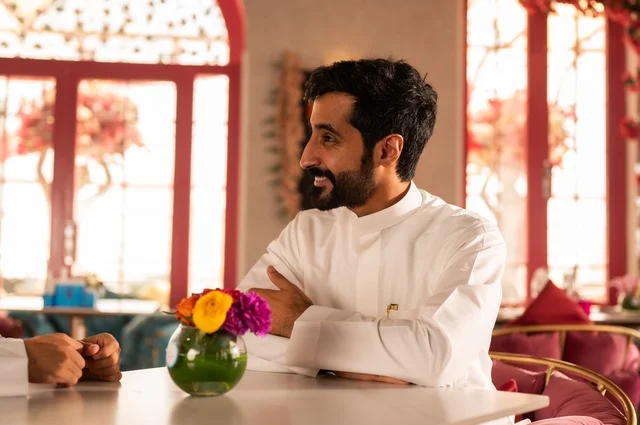 A side image of a young man in an upscale coffee shop, spending his free time sipping his favorite drink, enjoying cold beverages. An Arab Gulf Saudi man wearing a white thobe sits at a table in a café, showing gestures of happiness and joy.
