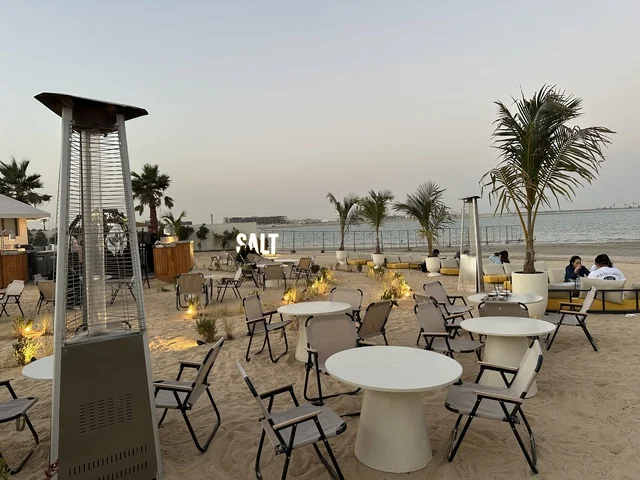 أشجار نخيل أضواء صغيرة حول جذوعها، سولت البحر وجهة ترفيهية وشاطئية عائلية تقع على طريق الكورنيش في الخبر في المملكة العربية السعودية، تجديدات إضافية وتطويرات في 3  يناير 2026، شاطئ رملي مع وجود مظلات وأشجار نخيل