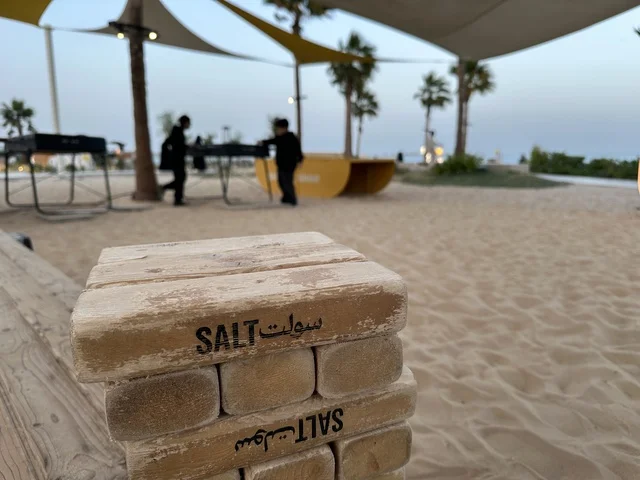 كتلة الخشبية في المقدمة تحمل اسم سولت،  شاطئ رملي مع وجود مظلات وأشجار نخيل، سولت البحر وجهة ترفيهية وشاطئية عائلية تقع على طريق الكورنيش في الخبر في المملكة العربية السعودية، تجديدات إضافية وتطويرات في 3  يناير 2026