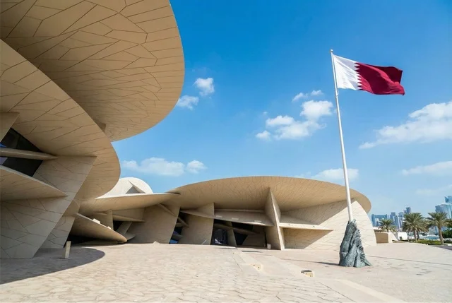 علم دولة قطر يرفرف في السماء الزرقاء, الاحتفال باليوم الوطني القطري, المتحف الوطني الإسلامي للفنون في العاصمة القطرية الدوحة, معالم سياحية مشهورة في وقت النهار، المباني الطينية على الطراز القديم