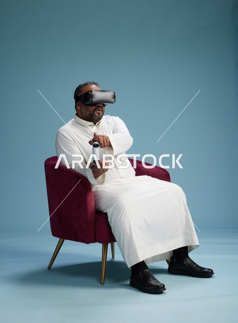 A portrait of a Saudi man wearing virtual reality glasses and holding a controller, enjoying video games, with a blue background.