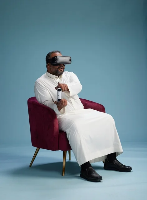 A portrait of a Saudi man wearing virtual reality glasses and holding a controller, enjoying video games, with a blue background.