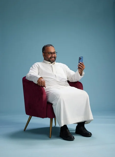 A smiling Saudi man wearing a white thobe during a video call on a mobile phone, selfie shot, blue background.