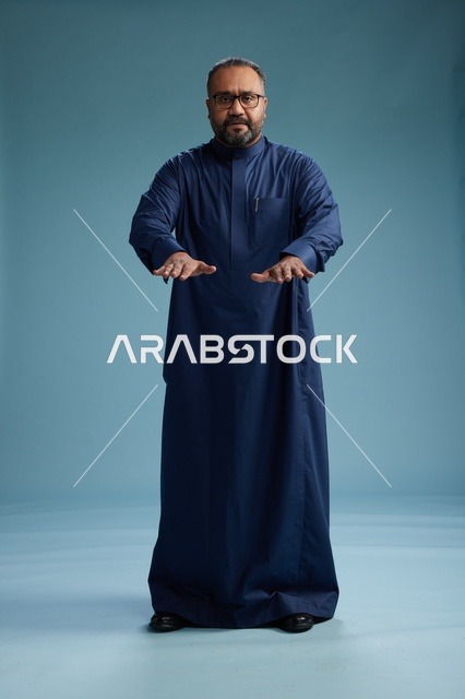 بورتريه لرجل سعودي يقف مرتدياً ثوب كحلي ينظر الى الكاميرا, كامل الجسم, خلفيه زرقاء