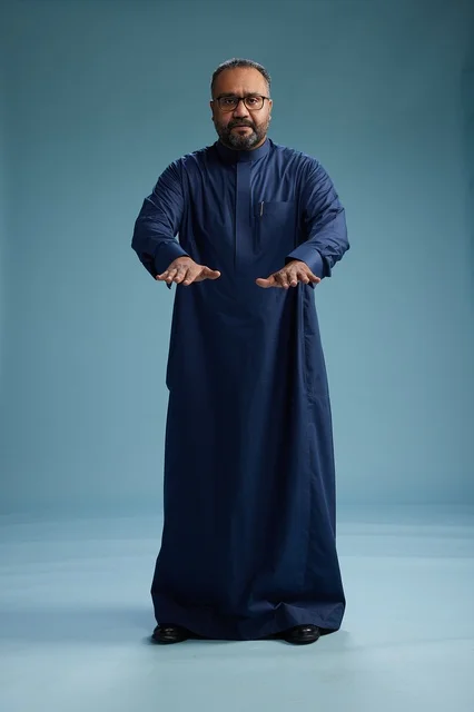 A portrait of a Saudi man standing wearing a navy blue thobe looking at the camera, full body, with a blue background.