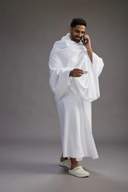 بورتريه لرجل سعودي بملابس الإحرام يحمل بطاقة بيضاء فارغه, مفهموم الحج والعمره, كامل الجسم, خلفيه رماديه