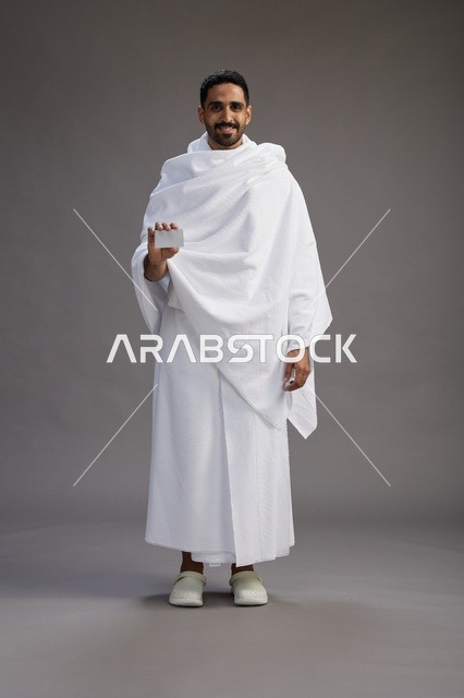 بورتريه لرجل سعودي بملابس الإحرام يحمل بطاقة بيضاء فارغه, مفهموم الحج والعمره, كامل الجسم, خلفيه رماديه