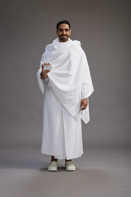 A portrait of a Saudi man in Ihram clothing holding a blank white card, representing the concepts of Hajj and Umrah, full body, gray background.