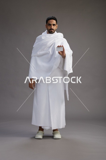 بورتريه لرجل سعودي بملابس الإحرام يحمل بطاقة بيضاء فارغه, مفهموم الحج والعمره, كامل الجسم, خلفيه رماديه