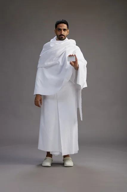 A portrait of a Saudi man in Ihram clothing holding a blank white card, representing the concepts of Hajj and Umrah, full body, gray background.