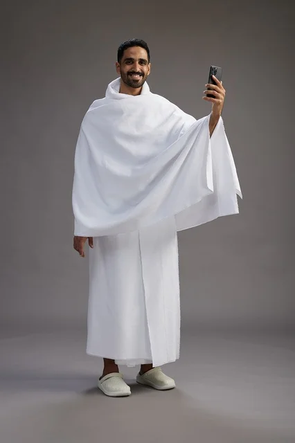 A portrait of a Saudi man in Ihram clothing smiling and taking a selfie, using a mobile phone, during Hajj and Umrah in Mecca, full body, gray background.